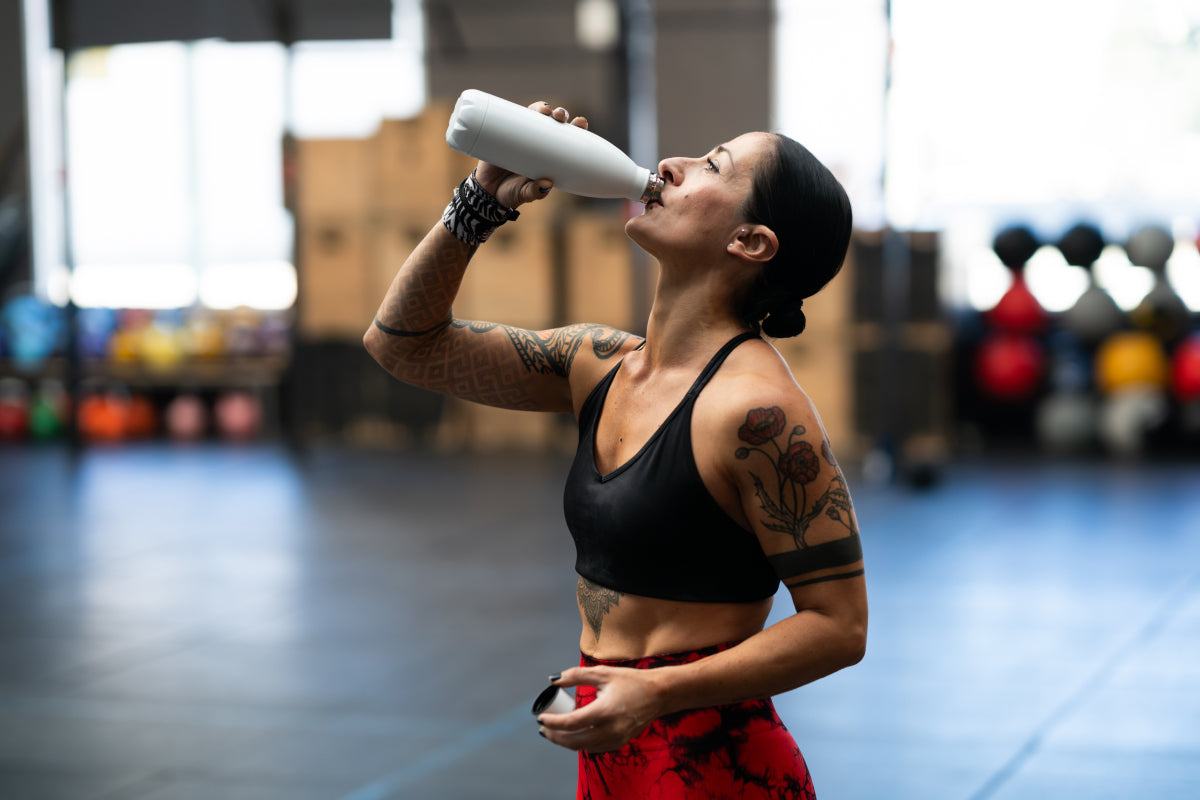 Eine fitte Frau mit Tattoos auf den Armen trinkt aus einer Wasserflasche in einem Fitnessstudio