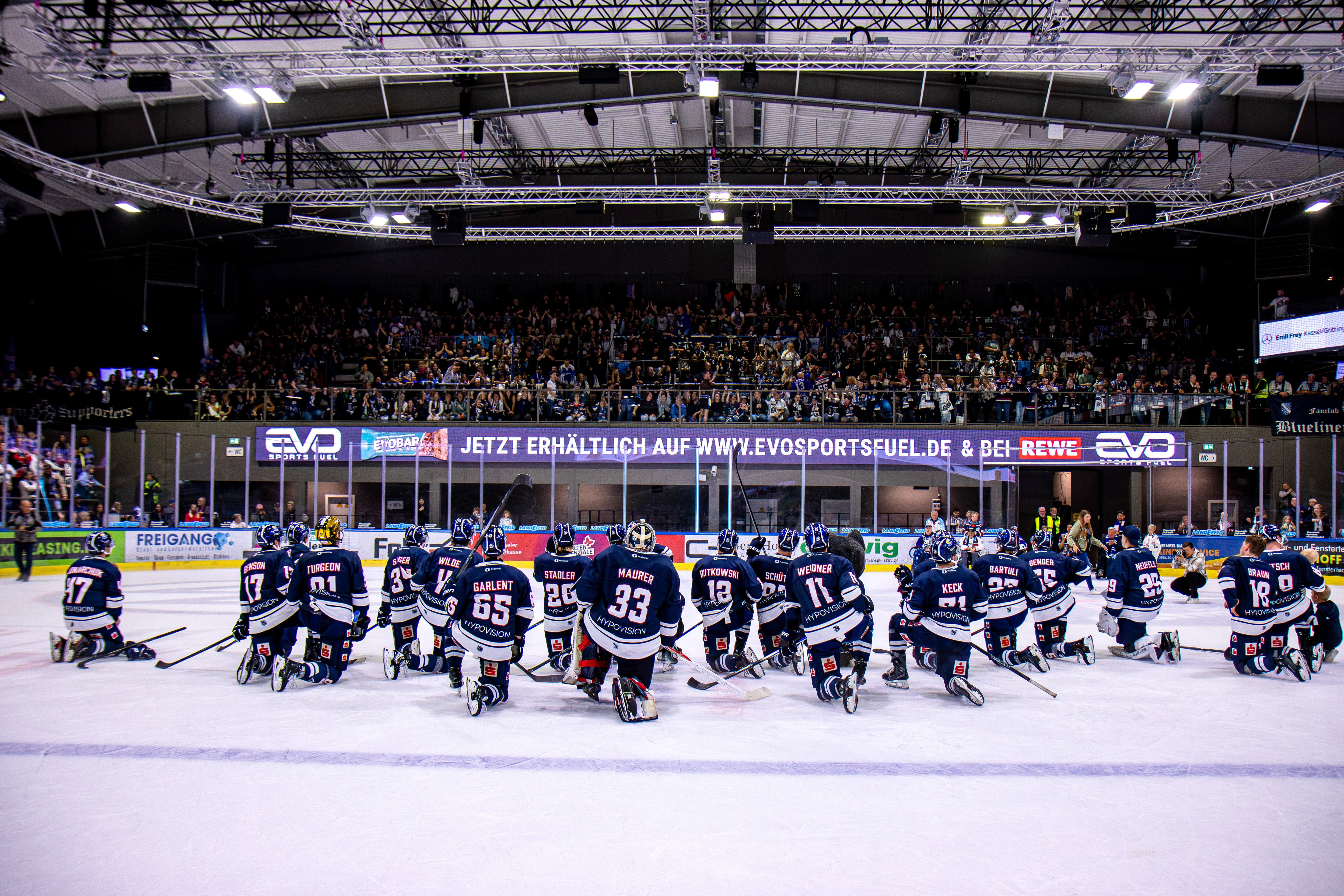 EVO SPORTS FUEL als Partner im Profisport: Eishockeyteam Kasseler Huskies kniet nach dem Spiel auf dem Eis vor voller Tribüne und bedankt sich bei den Fans im Stadion. Auf der Banderole evosportsfuel.de
